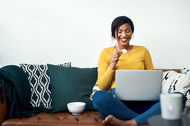 woman sitting on her sofa and eating potato crisps while using her laptop