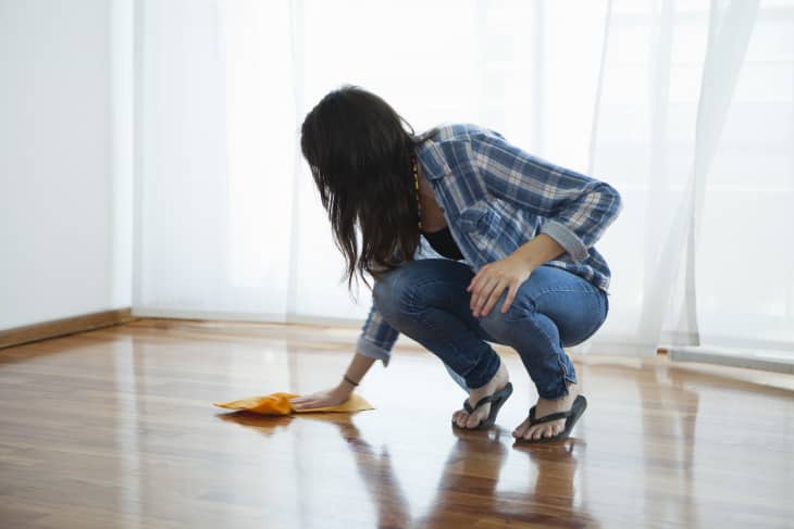 Woman wiping hardwood floor with window and sheer white curtains in background