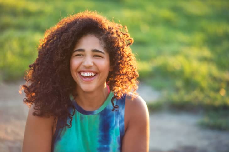 Smiling woman with curly brown hair wearing tie dyed tank top with green grass behind her