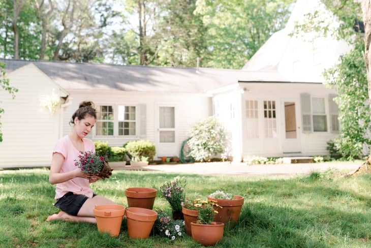 Woman planting flowers in terracotta pots on a grassy lawn in front of a white house.