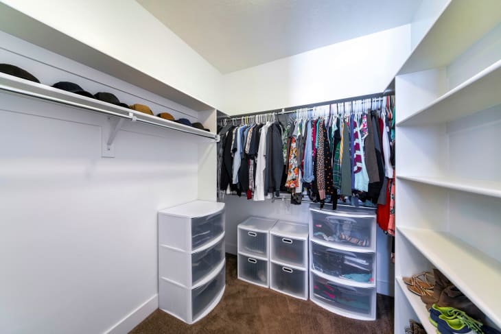 Walk-in closet with plastic wardrobe storage and wall mounted shelves and rods. There are caps on the top shelf, clothes hanging on a closet rod, and footwears on the lower shelves on the right.