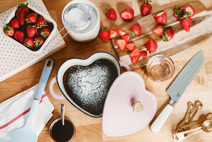Heart-shaped chocolate cake dusted with powdered sugar, surrounded by fresh strawberries, a knife, and baking tools on a wooden surface.