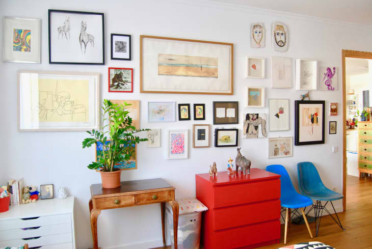 Gallery wall with diverse framed art, a wooden table with a potted plant, red drawers, and two blue chairs.