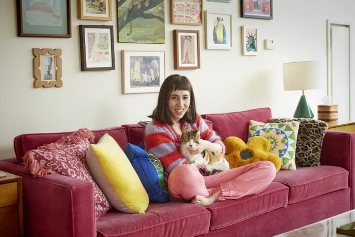 Woman sitting on a red sofa with colorful pillows, holding a cat, surrounded by framed art on the wall.