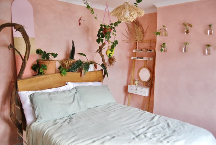 Bedroom with peach/pink walls, plants. Bed has mint green bedding, wood headboard
