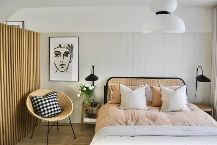 bedroom with white slatted walls, natural fiber rug, bed with rattan headboard and pale terracotta and white bedding, modern art, framed, straw/wicker accent chair