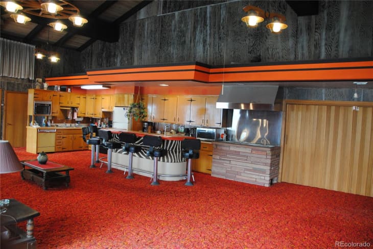 Retro kitchen with orange accents, wood cabinets, red carpet, bar stools, and stone fireplace.