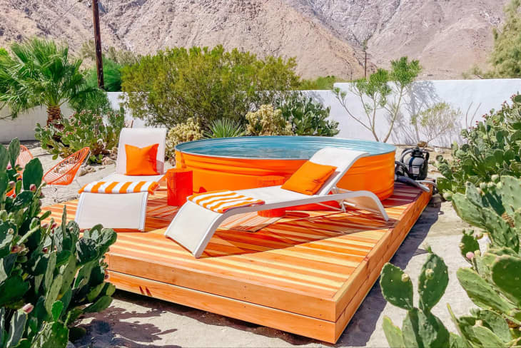 Palm Springs raised wood patio/deck with Dani Dazey designed orange stock tank pool, white lounge chairs with orange and white striped towels and orange pillows, cactus around, mountains in the background