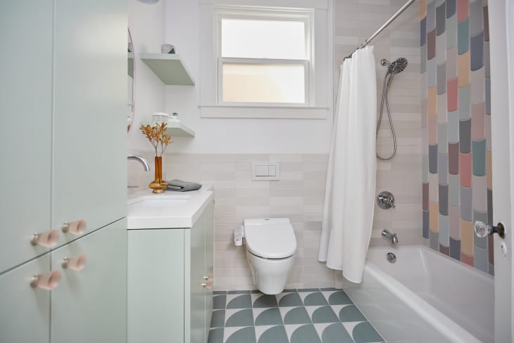 pastels, light green cabinets and vanity, dark sage green arched square tile with white corner, pastel colorful tile wall in tub, beige tile half wall behind toilet, floating shelves, white shower curtain, tub shower combo, floating toilet, orange vase with small flowers