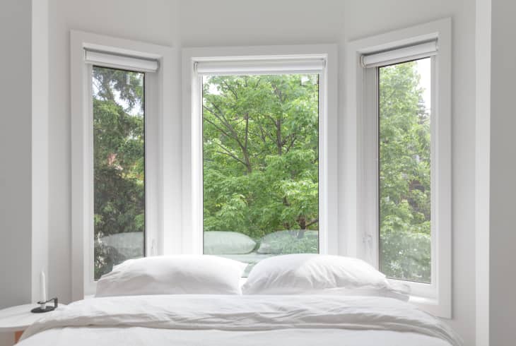 Bed with white linens in front of three large windows overlooking lush green trees.