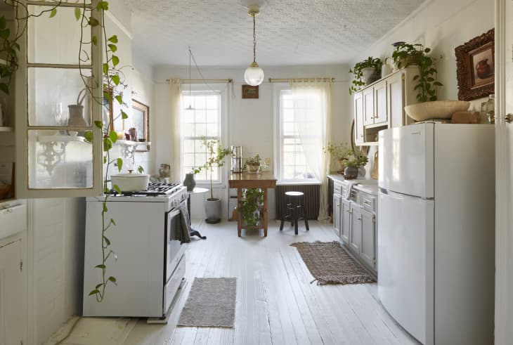 Bright kitchen with white cabinets, hanging plants, a wooden table, and a white fridge.