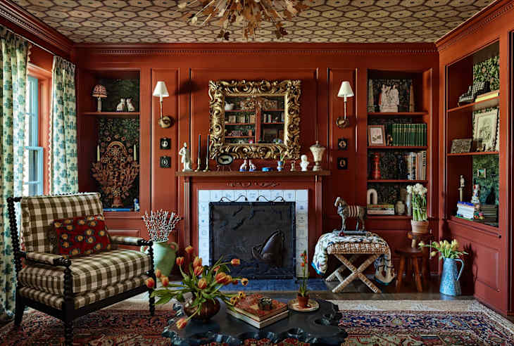 Cozy living room with red walls, a patterned ceiling, a fireplace, and a checkered armchair, adorned with books and decor.
