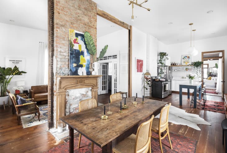 Open-concept living and dining area with rustic wooden table, brick fireplace, abstract art, and modern kitchen.