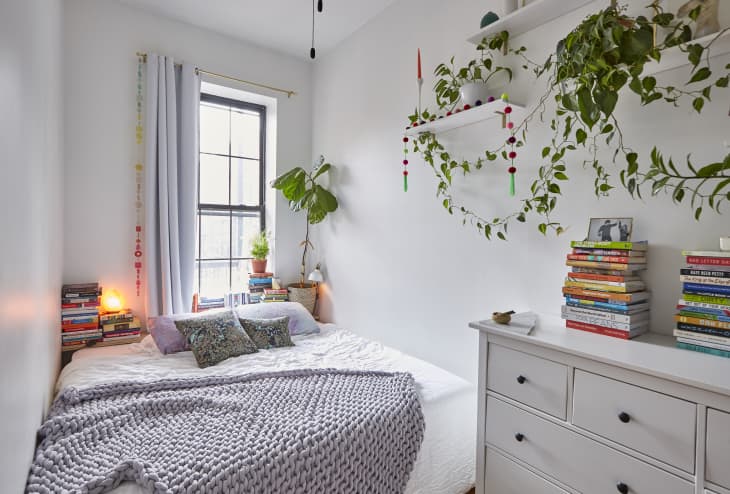 Cozy bedroom with a bed, gray knit blanket, books, potted plants, and hanging greenery on white walls.