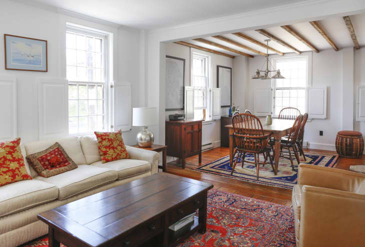 Living room with cream sofa, red patterned cushions, wooden coffee table, and dining area with wooden chairs and blue rug.