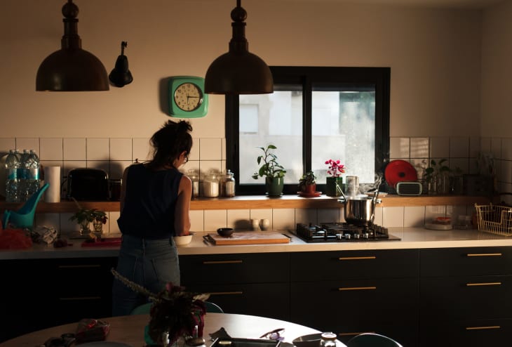 Woman cooking in a dimly lit kitchen with plants on the windowsill, a clock on the wall, and a pot on the stove.