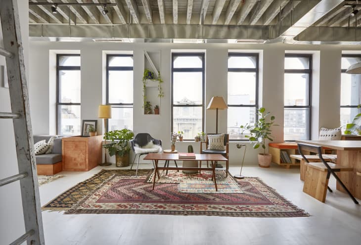 Loft living room with large windows, eclectic rugs, wooden furniture, plants, and modern decor.