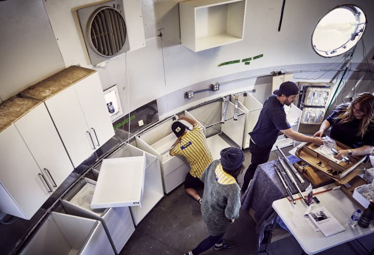 People assembling white kitchen cabinets in a circular room with a round window and various tools on a table.
