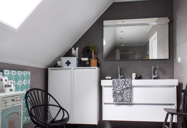 Bathroom with sloped ceiling, large mirror, double sink, white cabinets, black chair, and patterned towel.