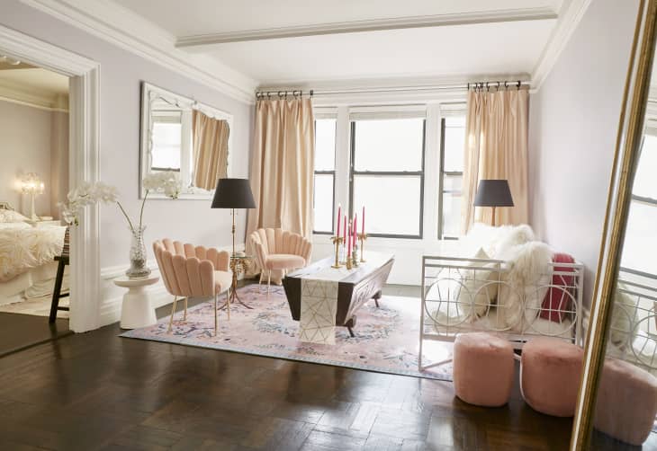 Elegant living room with pink velvet chairs, floral rug, mirrored coffee table, and large windows with peach curtains.