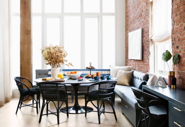 Dining area with black chairs, round table, dried flowers, and a brick wall. Large windows and a cozy bench with cushions.