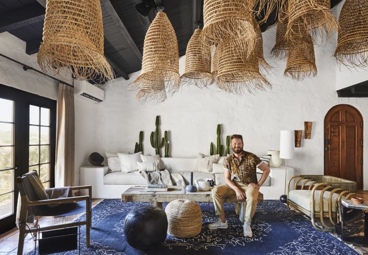 Living room with wicker pendant lights, white sofa, cactus decor, and a man sitting on a wooden chair, blue patterned rug.
