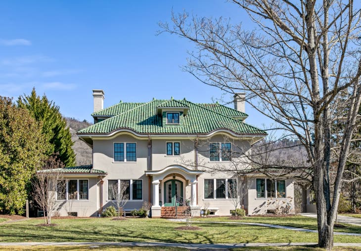 Two-story house with green tile roof, arched entryway, and large windows, surrounded by trees and a lawn.