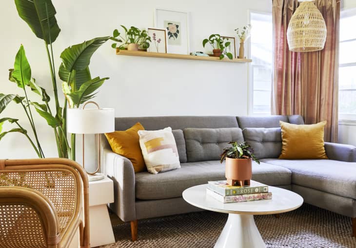 White living room with gray sectional sofa, pink drapery, and boho accents like plants and a cane chair and woven light fixture