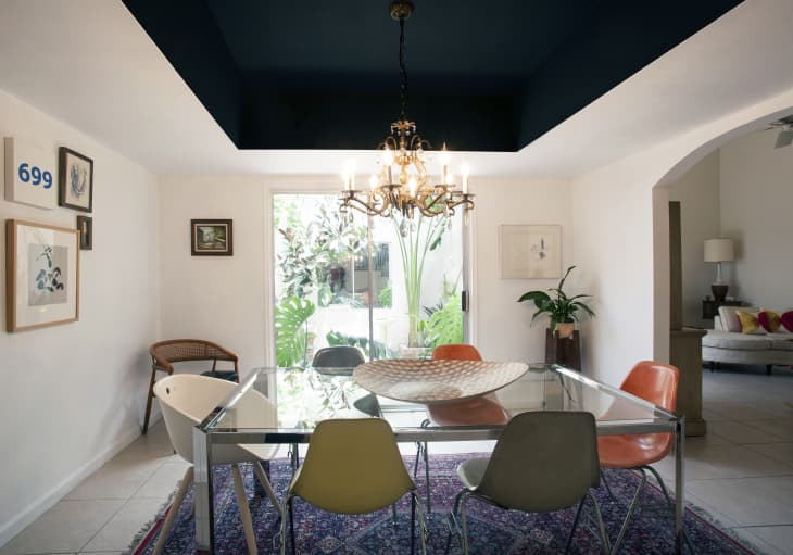 Dining room with glass table, colorful chairs, chandelier, and wall art, overlooking a garden through large windows.