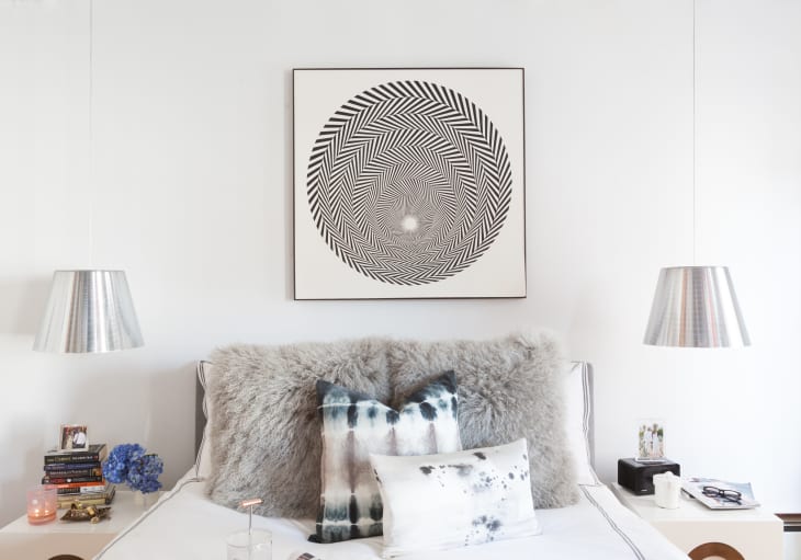 Bedroom with abstract art above bed, fluffy gray throw, tie-dye pillows, and silver pendant lamps.