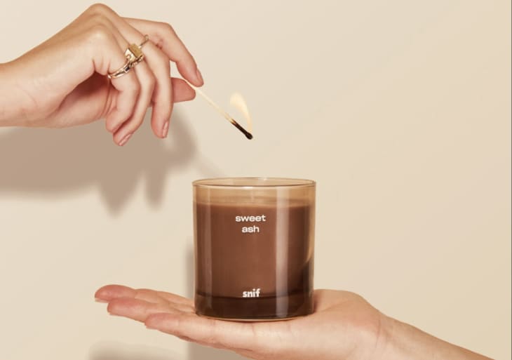 Hand lighting a "sweet ash" candle in a glass jar held by another hand.