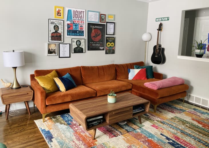 Living room with orange velvet sofa, mid-century tables, colorful rug, and concert artwork on wall