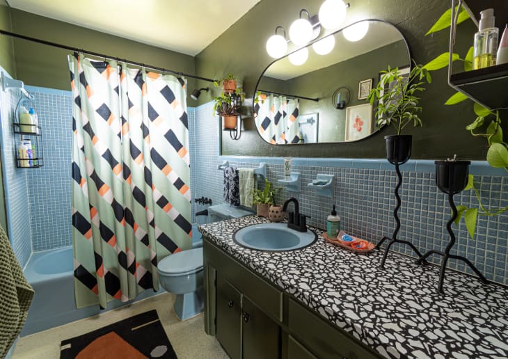 Retro bathroom with blue tiles, patterned shower curtain, terrazzo countertop, oval mirror, and plants on shelves.