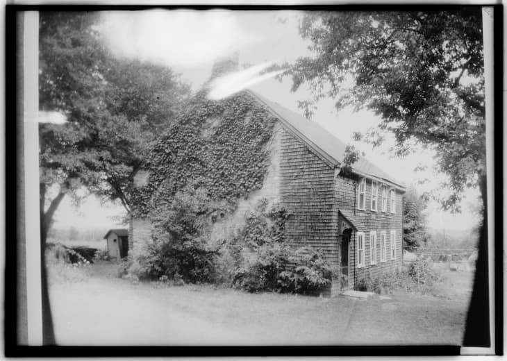 Historic house with ivy-covered walls, multiple windows, and a small outbuilding surrounded by trees and grass.