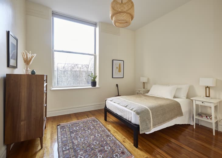 Minimalist bedroom with a wooden dresser, large window, woven pendant light, and a bed with beige bedding.