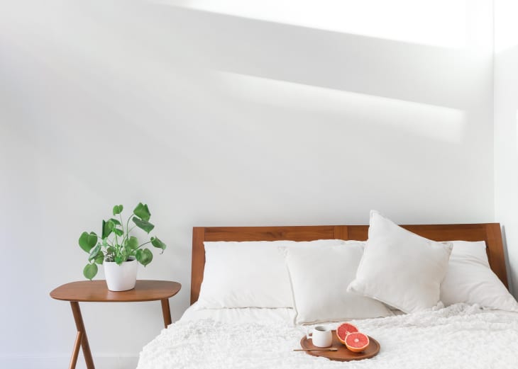 Bed with white pillows and blanket, wooden headboard, side table with plant, and tray with grapefruit and cup.
