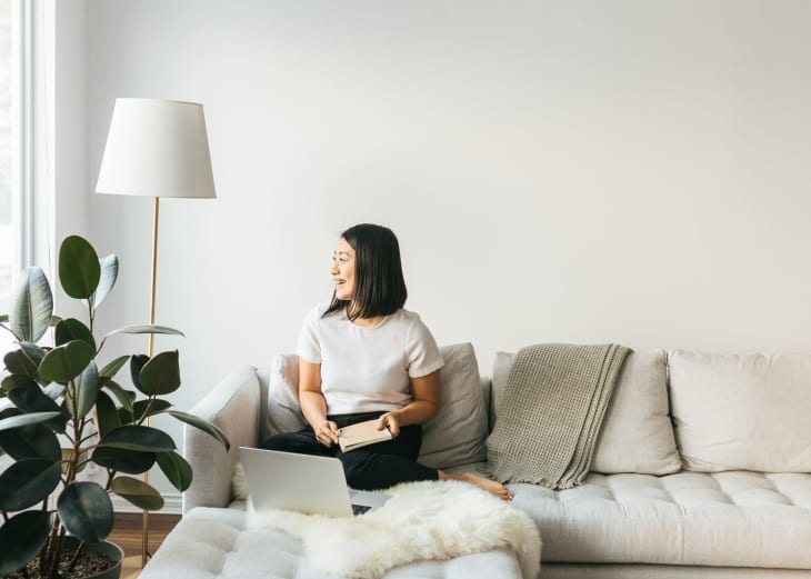 Woman sitting on a cream sofa with a book, next to a large plant and floor lamp, looking out the window.