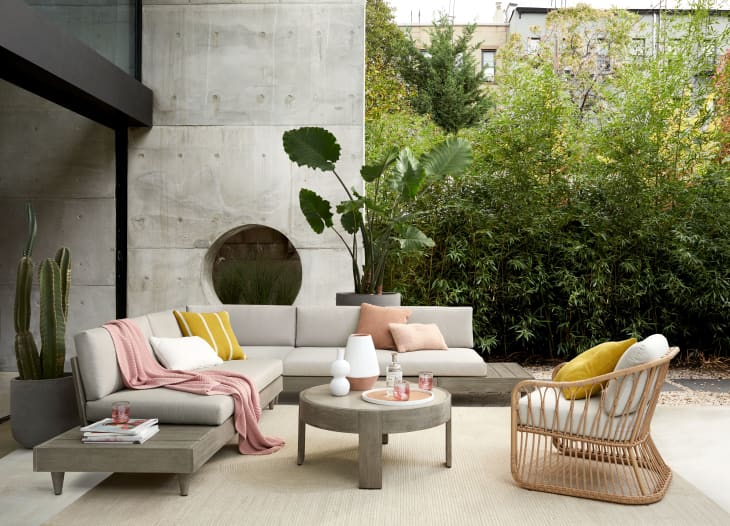 Outdoor patio with beige sectional sofa, pink and yellow cushions, round coffee table, and wicker chair, surrounded by greenery.