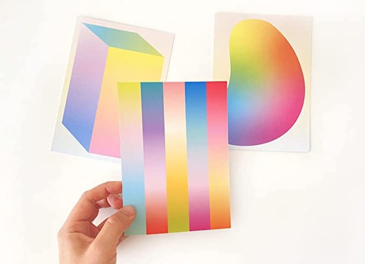 Hand holding a colorful gradient postcard with two abstract gradient art prints in the background.