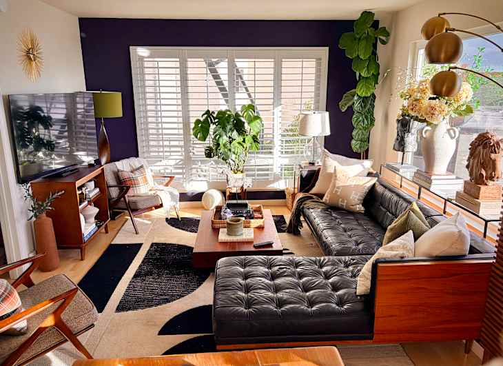 Modern living room with a black leather sectional, wooden coffee table, decorative pillows, and large plants.