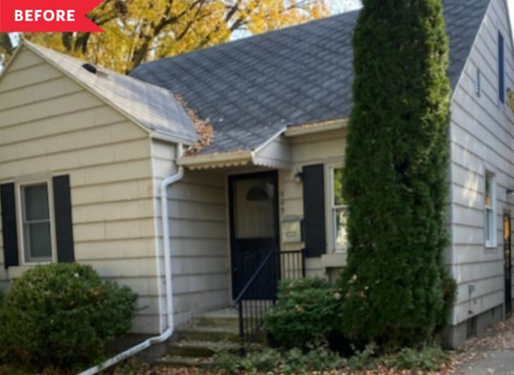 Small beige house with dark shutters, a sloped roof, and a tall evergreen tree in front.
