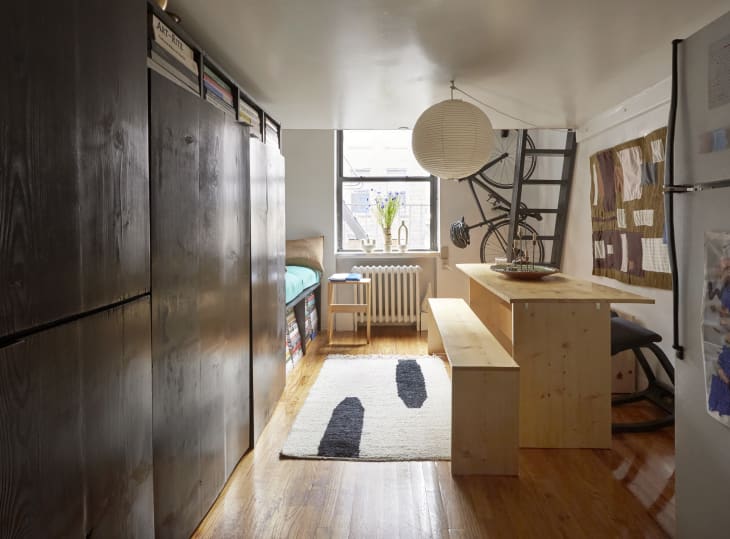 Wide angle of a small studio apartment with a long wall of black storage cabinets to the left and a natural wood dining table and bench