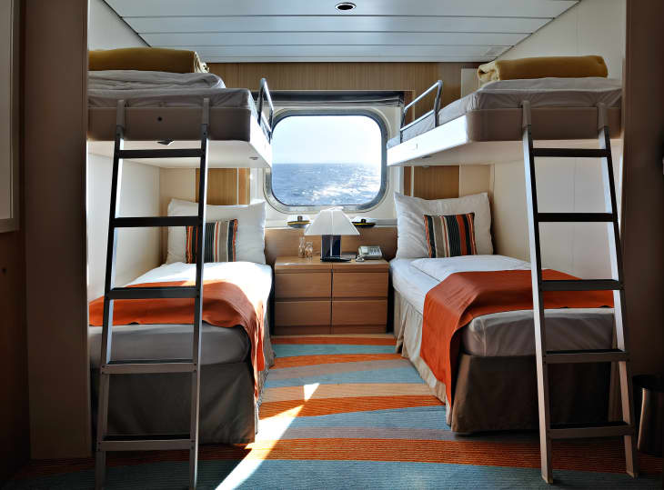 Cruise ship cabin with bunk beds, orange accents, striped pillows, and a window view of the ocean.
