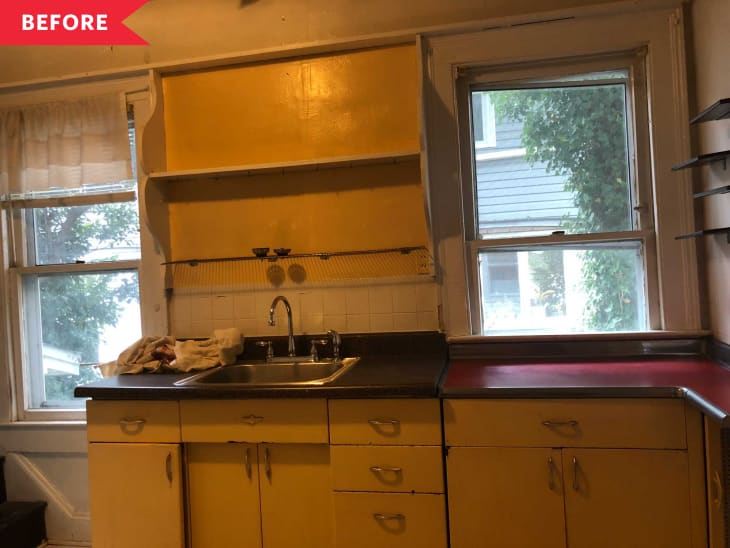 Yellow kitchen cabinets with a stainless steel sink, red countertop, and two windows with white curtains.
