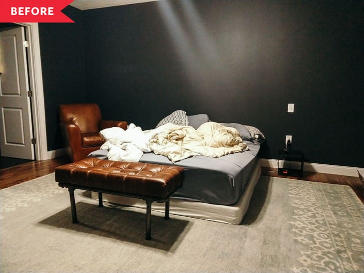Messy bed in a dark-walled room with a brown leather chair, tufted bench, and patterned rug.