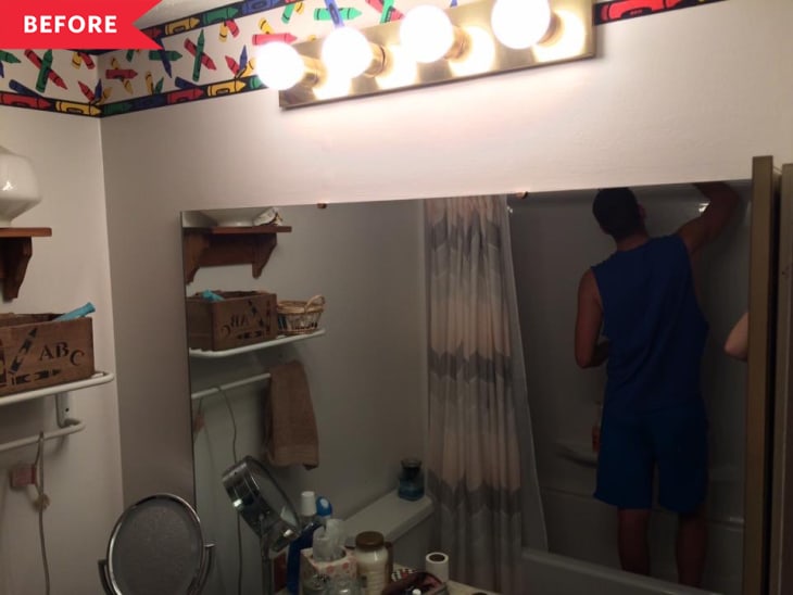 Man in blue outfit cleaning bathtub in cluttered bathroom with bright vanity lights and colorful wallpaper border.
