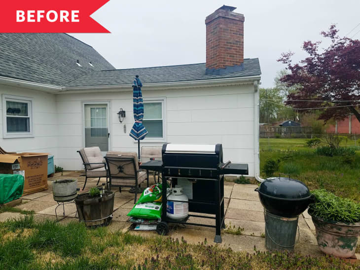 Patio with grill, potted plants, outdoor chairs, and a striped umbrella against a white house with a brick chimney.