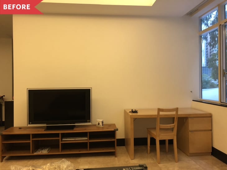 TV on wooden stand next to a light wood desk and chair, near a window.