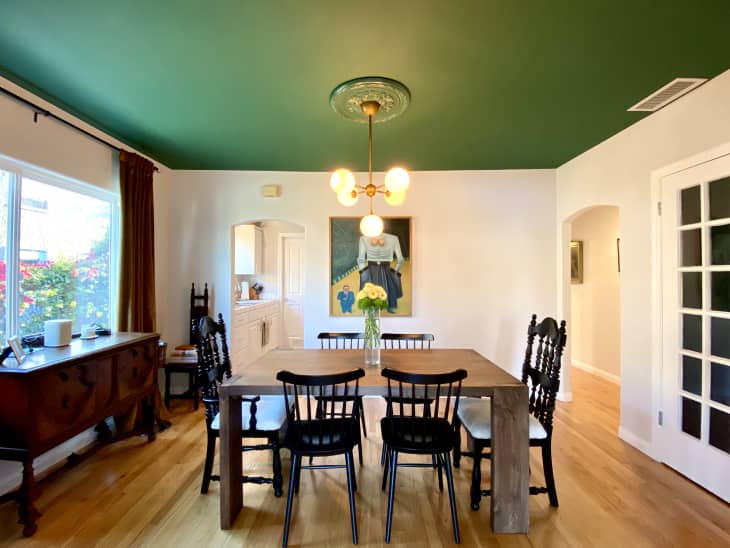 Dining room with green ceiling, wooden table, black chairs, chandelier, and a vase of yellow flowers.