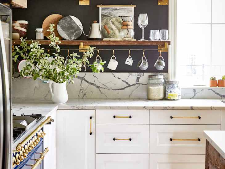 Kitchen with marble countertops, blue stove, hanging mugs, glassware, and a vase with greenery on a shelf.
